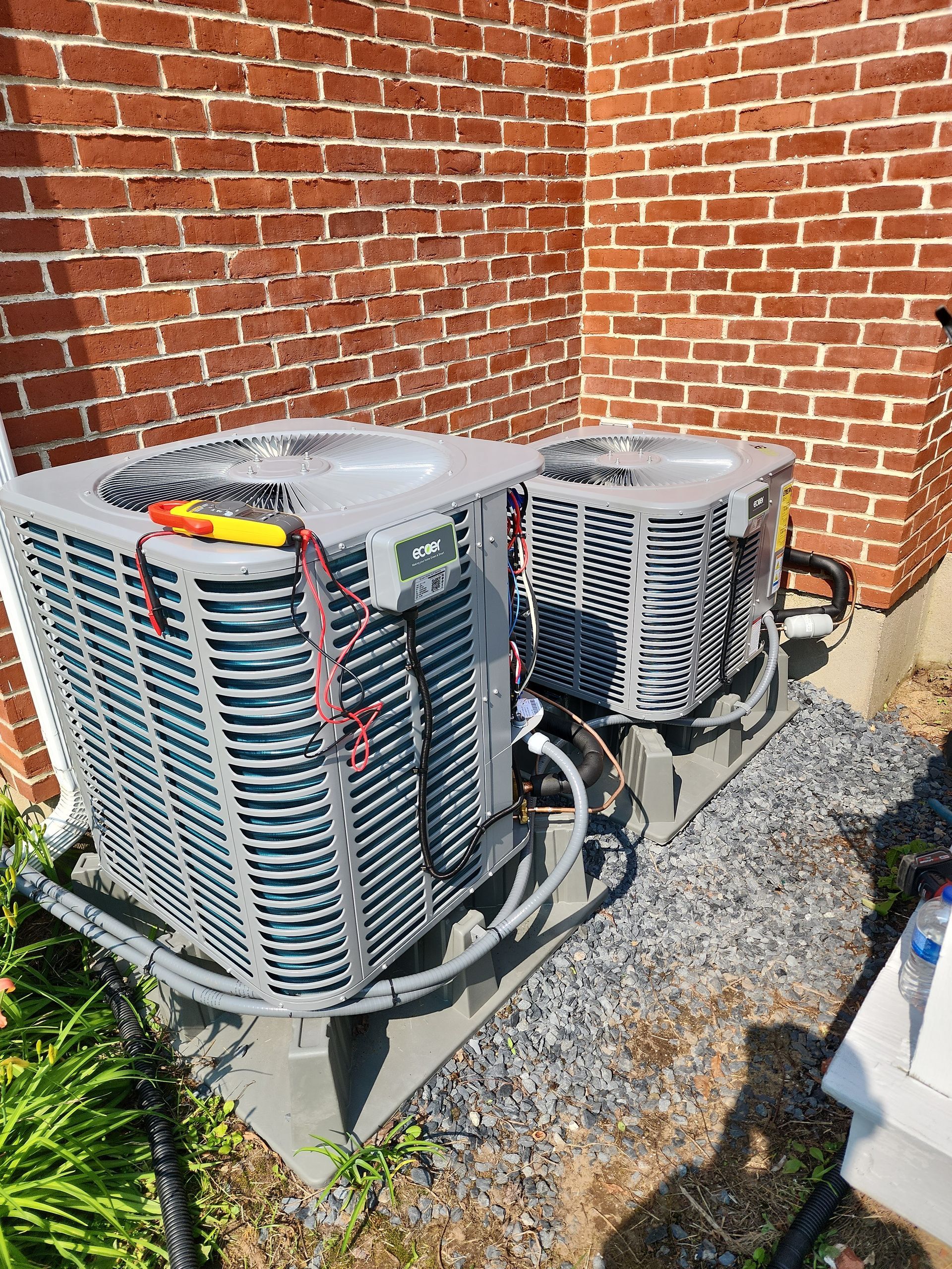 Two outdoor air conditioning units, gray and silver, sit on a concrete pad near a brick wall, with tools on one unit.