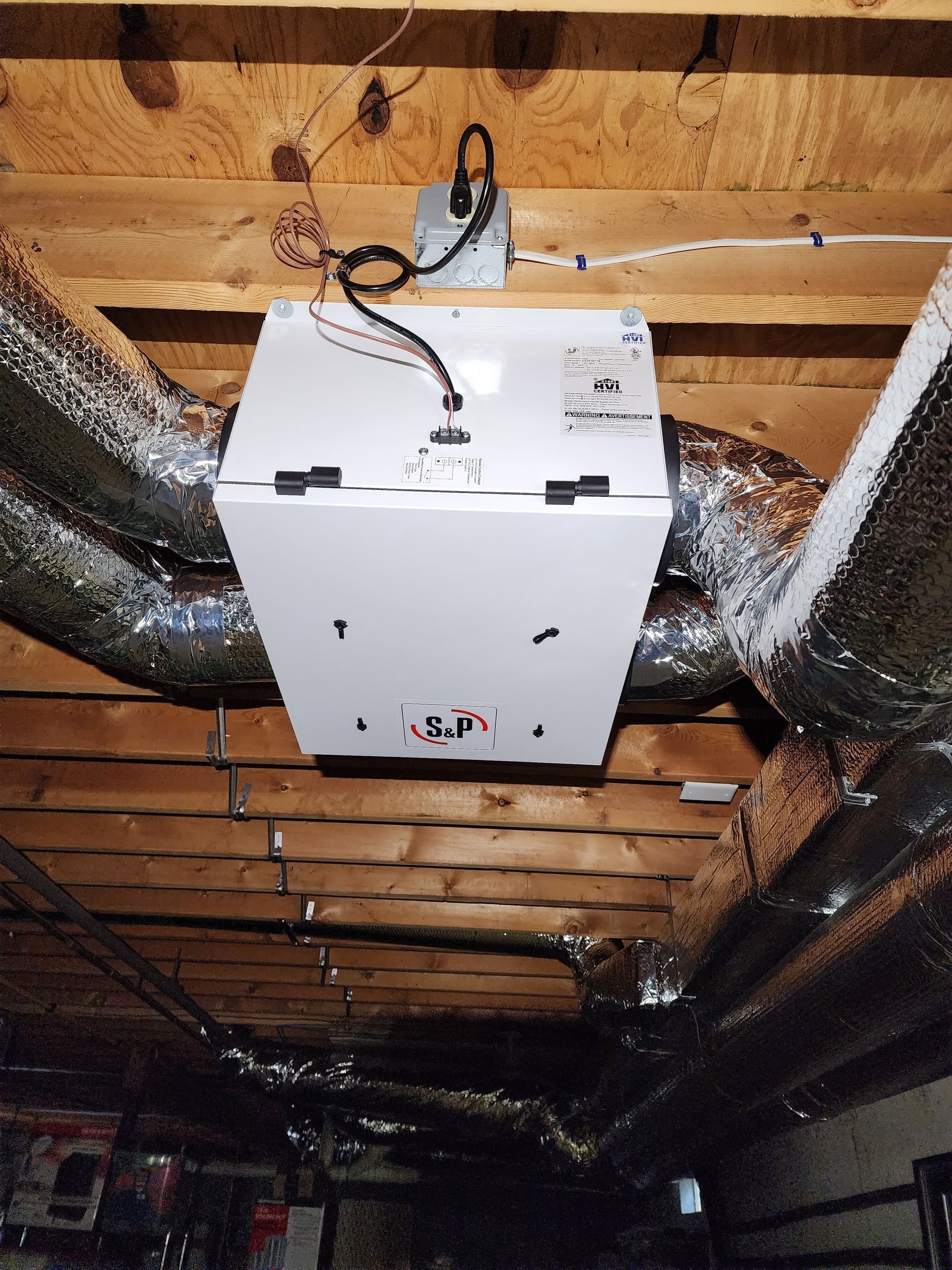 White ventilation system box suspended in a wooden ceiling, connected to ducts.