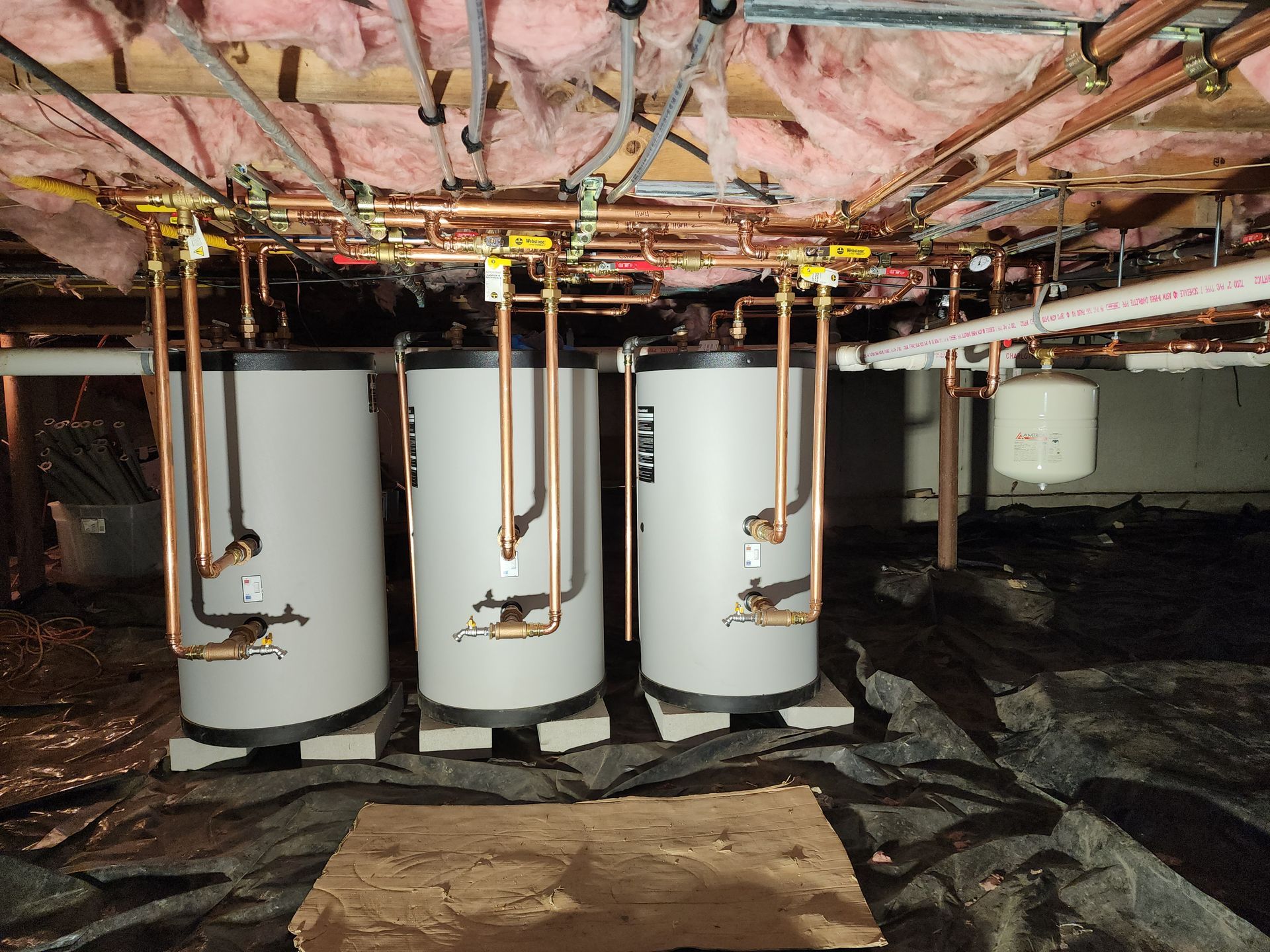 Three silver water heaters installed in a basement, copper pipes overhead, pink insulation.