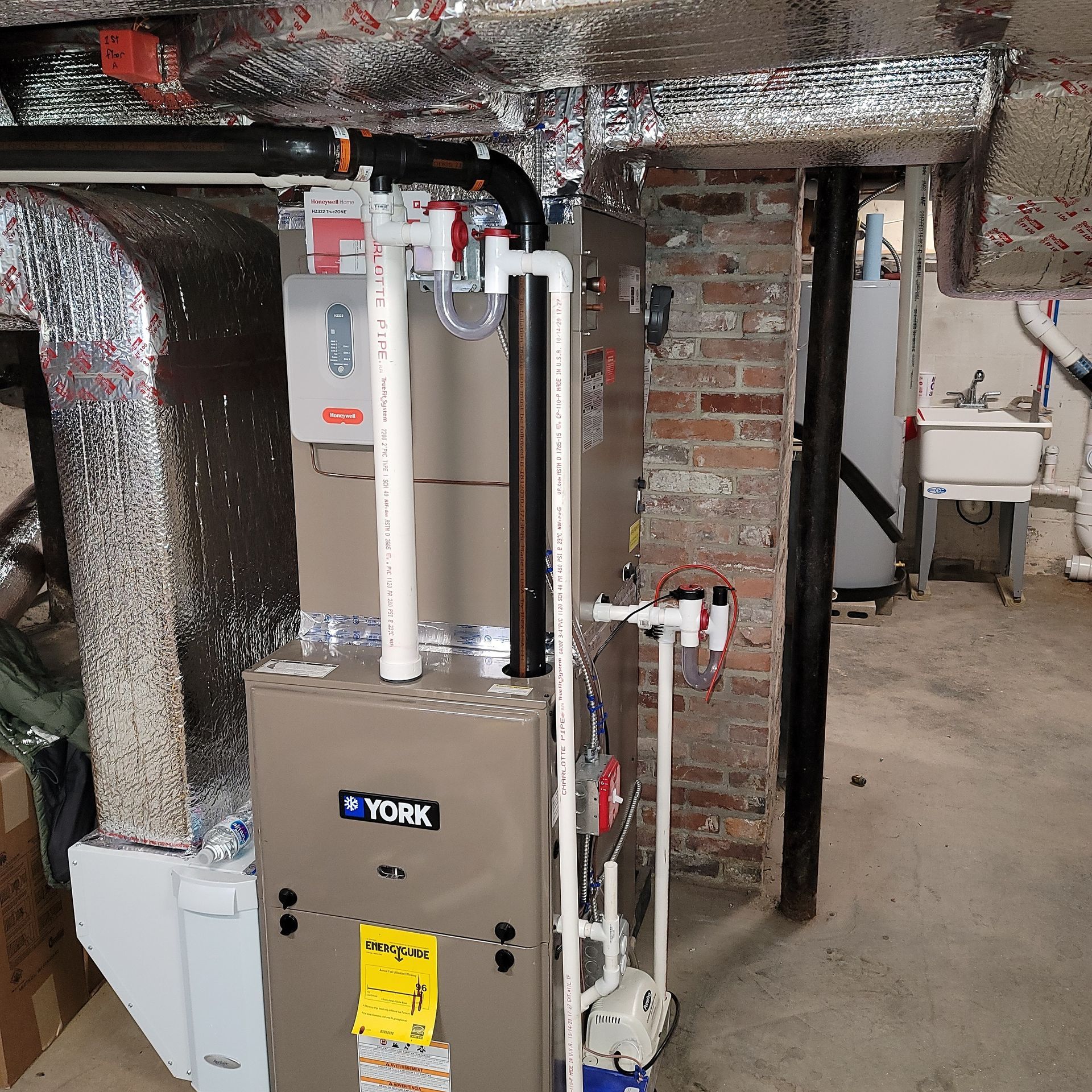 A furnace and connected pipes in a basement next to a brick wall and water basin.