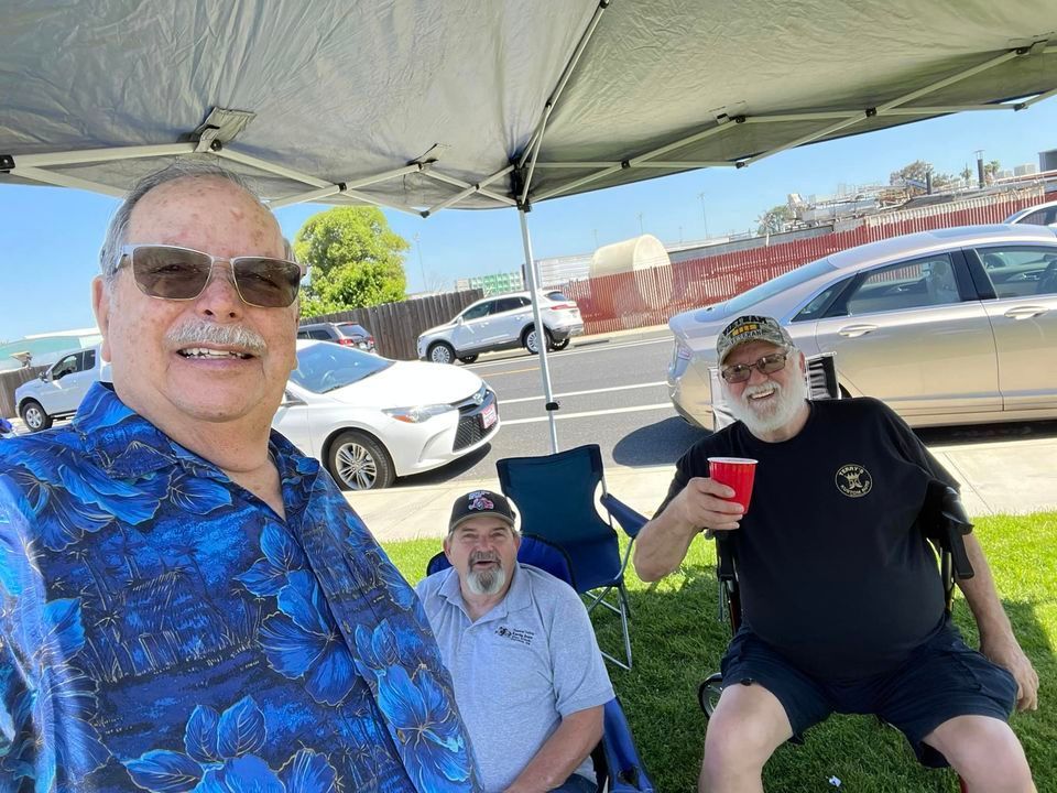 Three men are sitting under a tent in a parking lot.