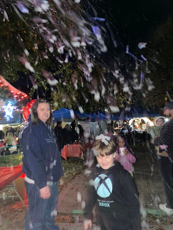 A man and a boy are standing under a tree with snow falling from it.