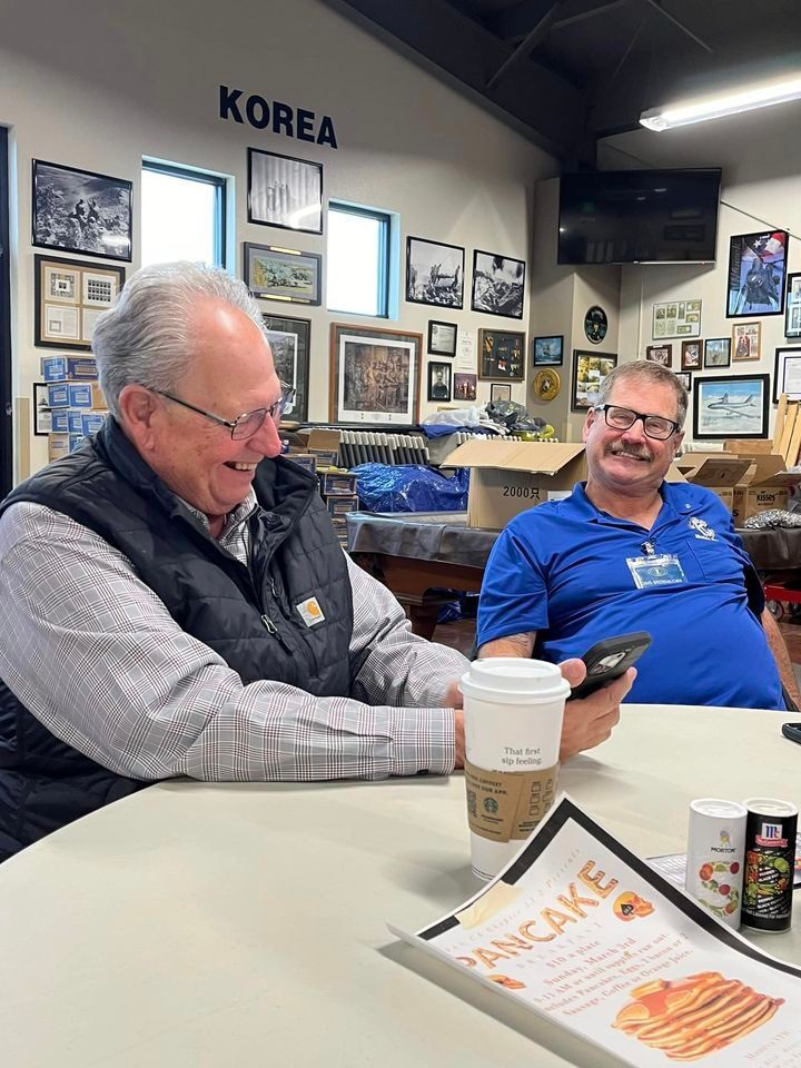 Two men are sitting at a table in a room with a sign that says korea on the wall.