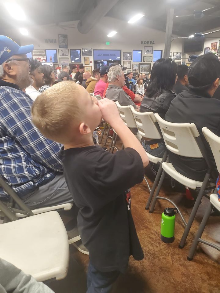 A young boy is drinking from a green water bottle while sitting in a crowded room.