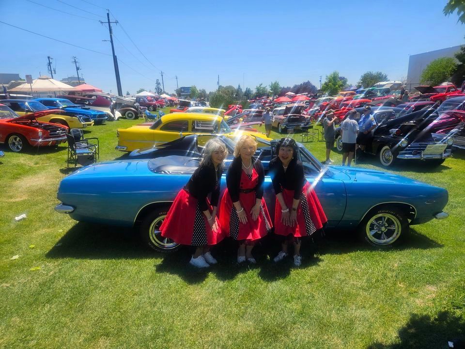 Three women are posing in front of a blue car at a car show.