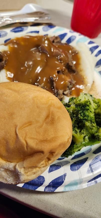 A paper plate topped with a hamburger , gravy , broccoli and mashed potatoes.