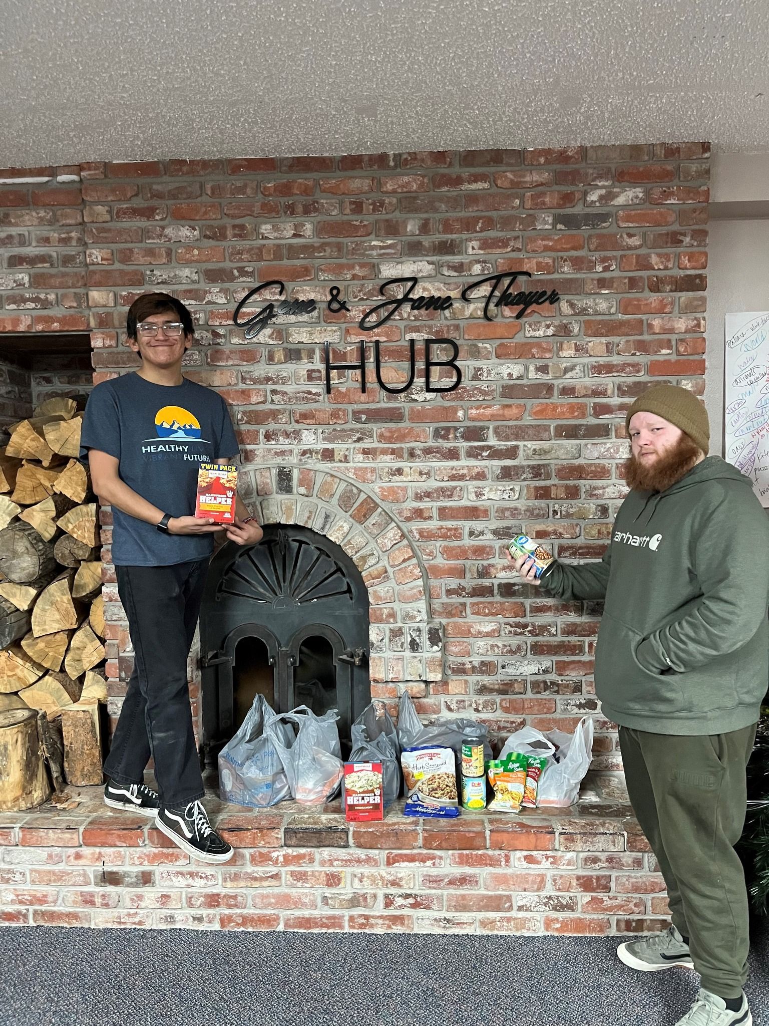 2 young adult males holding some food donations in front of a fireplace