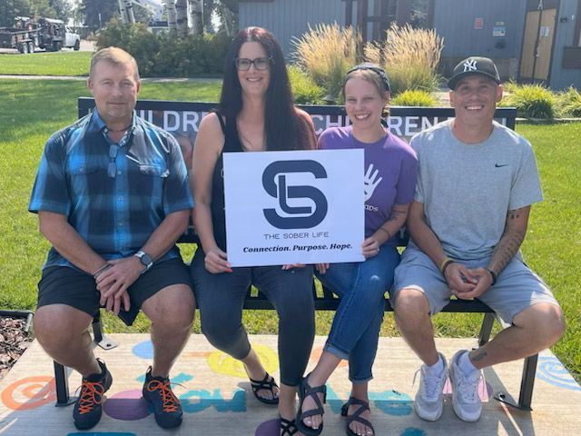 Four people sit on a park bench, holding a sign that reads