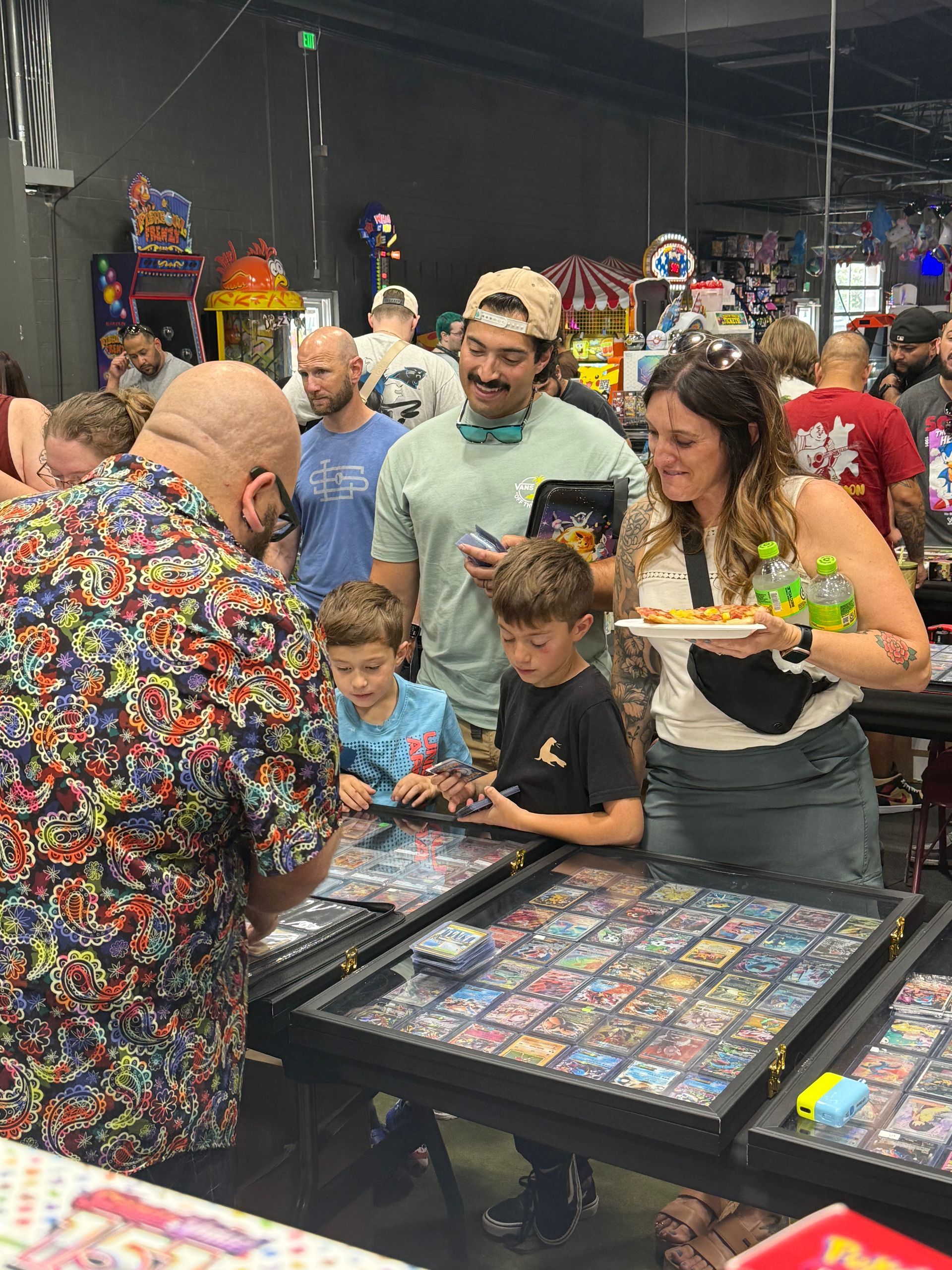 People browsing trading cards at a convention, including children.  A man in a colorful shirt is showing something to them.