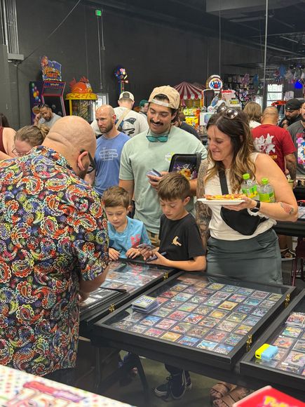 People browsing trading cards at a convention, including children. A man in a colorful shirt is showing something to them.