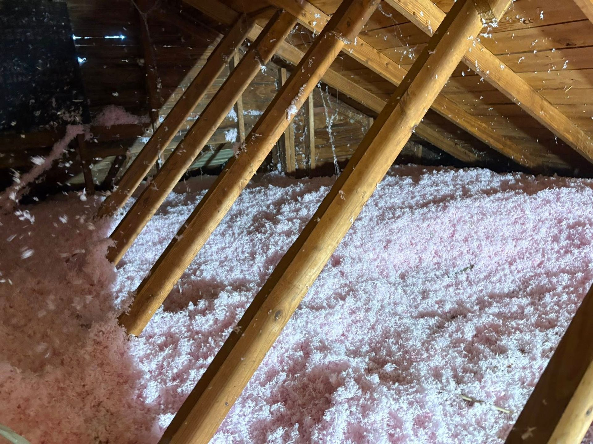 Wooden rafters above a snowy attic or crawlspace floor with pink insulation dusted in white snow