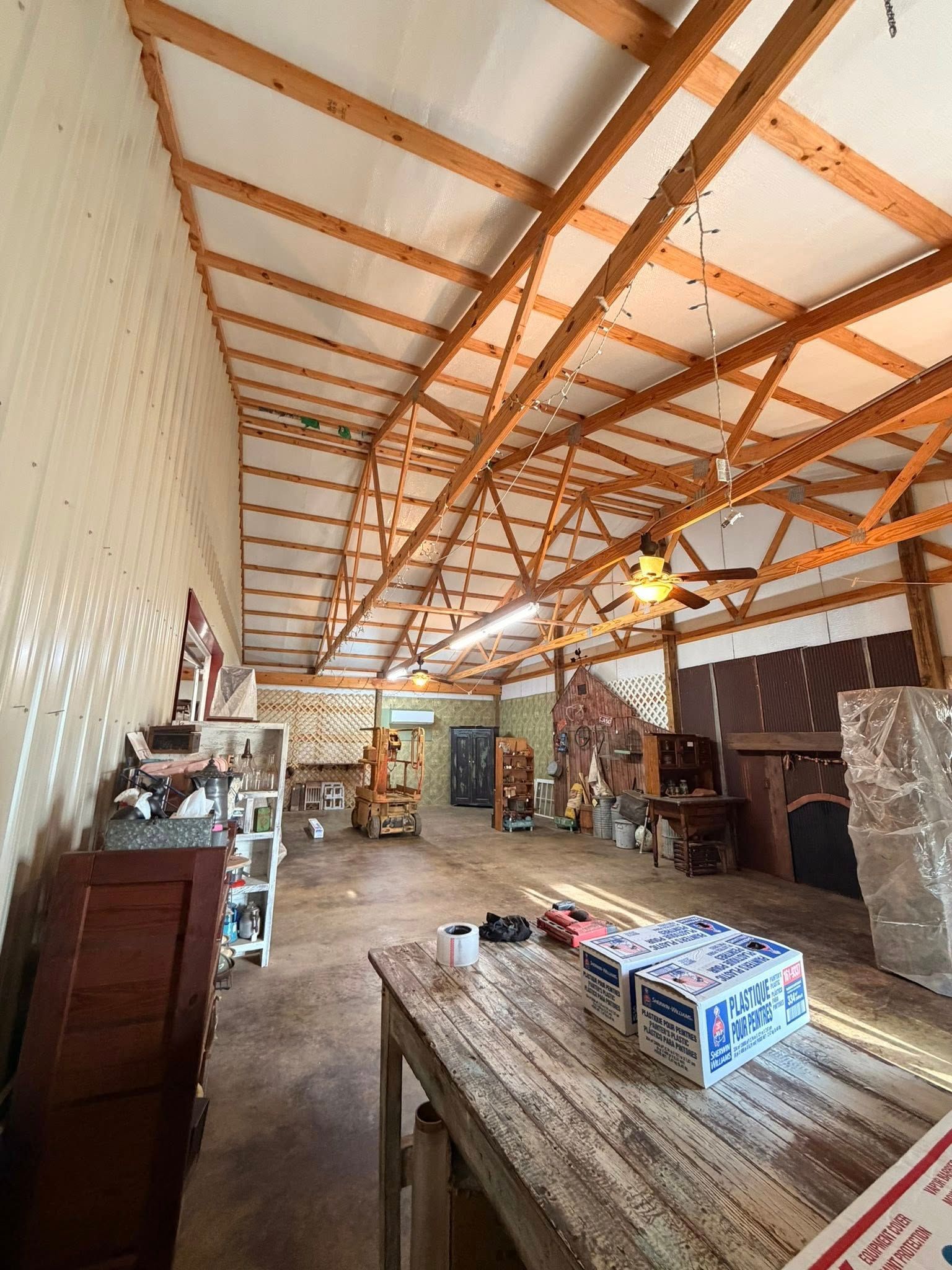 Long workshop interior with wood trusses, a workbench, shelving, and boxes under bright ceiling lights