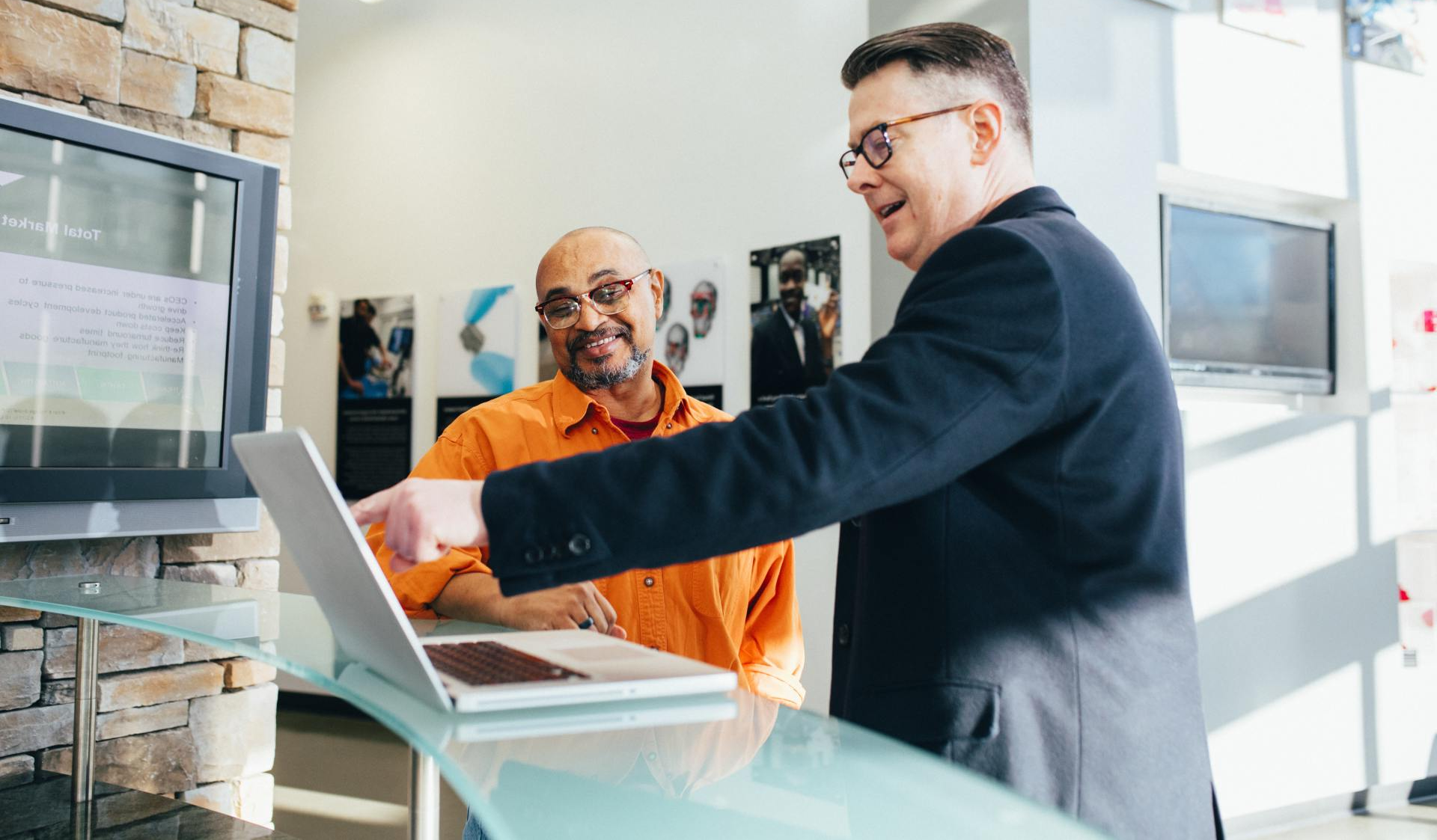A man is pointing at a laptop computer while another man looks on.