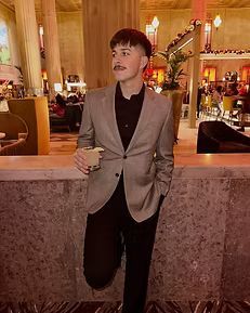 Man in blazer and black shirt holding a drink, leaning against a marble bar in a lit indoor setting.