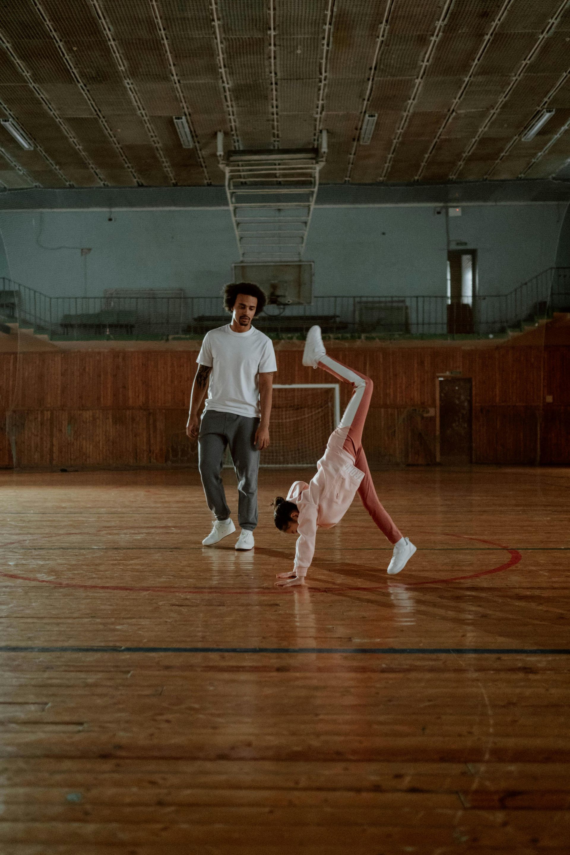 A man and a girl are doing a handstand on a wooden floor in a gym.