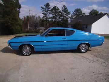 A bright blue vintage muscle car parked on a paved driveway in front of a house on a sunny day.