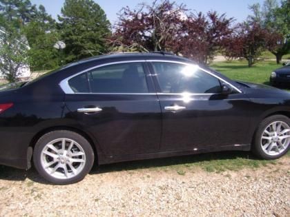A side view of a black sedan parked on a gravel surface outdoors with trees in the background.