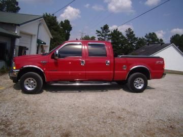 A bright red Ford crew cab pickup truck parked on a gravel lot in front of white buildings on a sunny day.