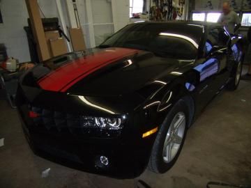 A black Chevrolet Camaro with a prominent red racing stripe on the hood, parked inside a garage.