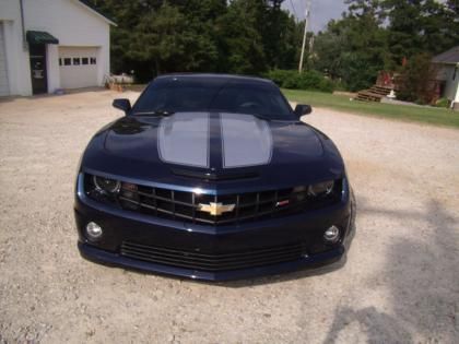 A dark blue Chevrolet Camaro with a silver center hood stripe, parked on a gravel driveway in front of a white building.