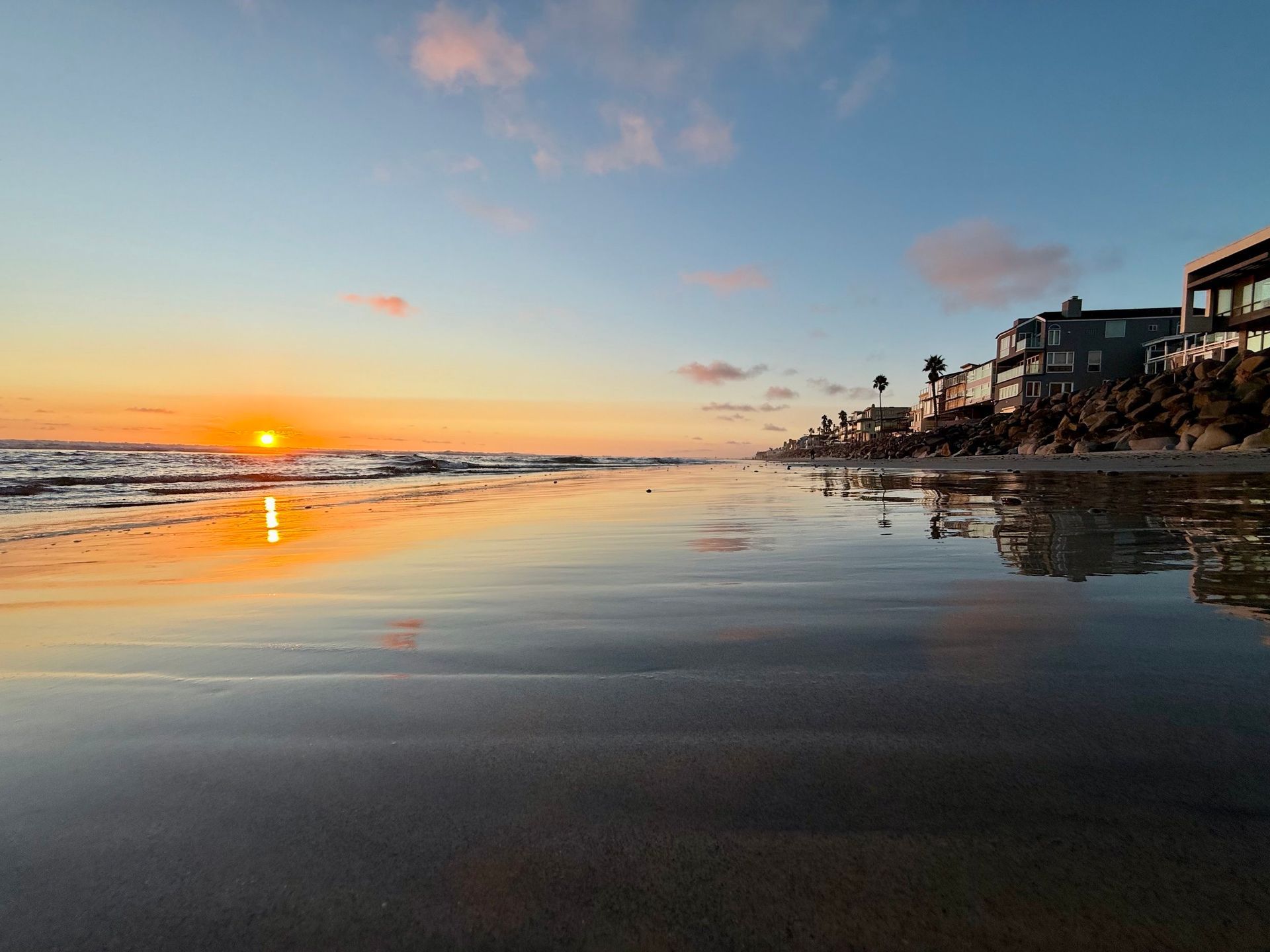 Sunset over a calm beach with wet sand reflecting orange sky and shoreline houses