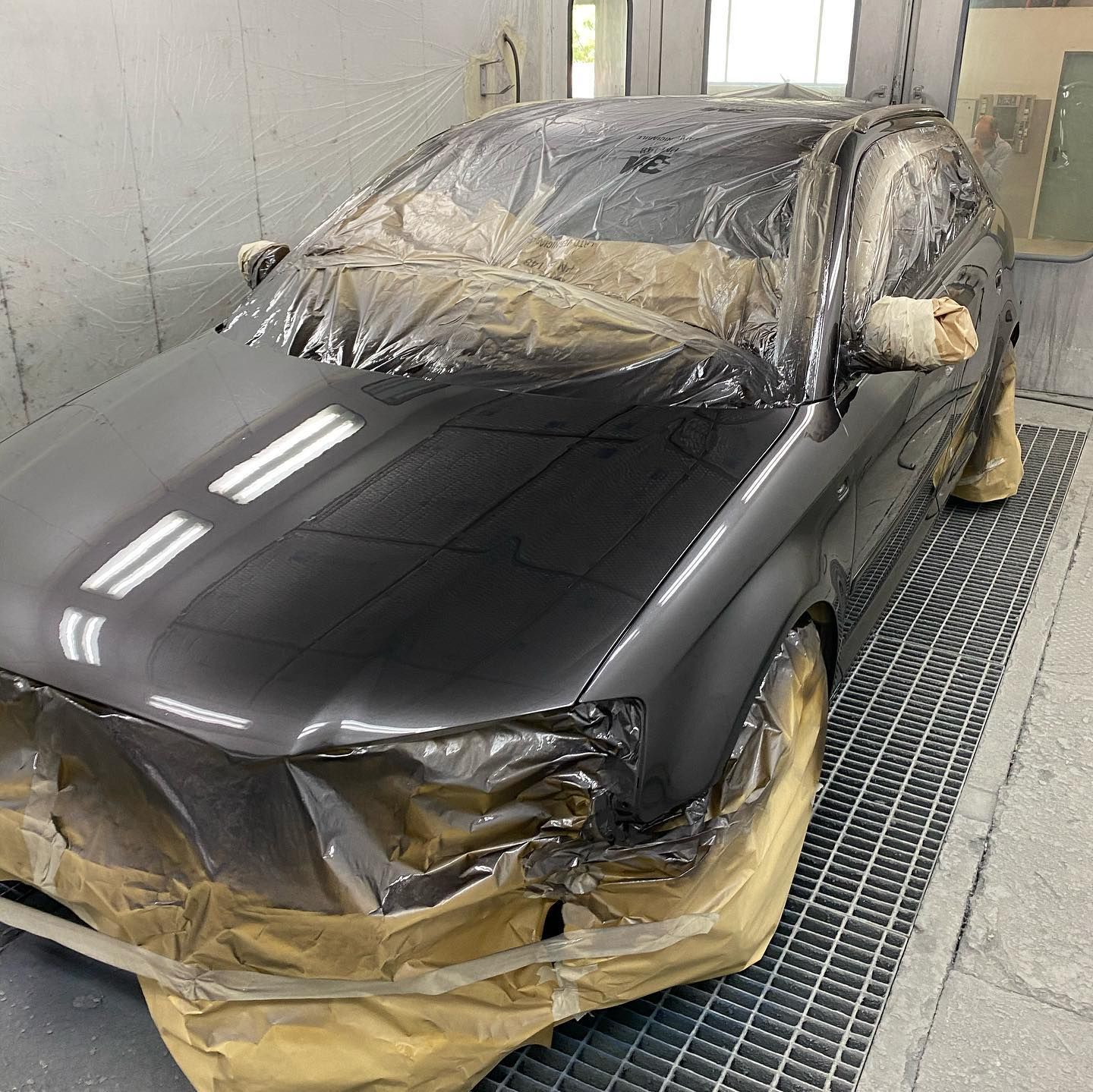 Un'auto grigio scuro in una cabina di verniciatura, ricoperta di plastica e nastro adesivo, pronta per essere verniciata.