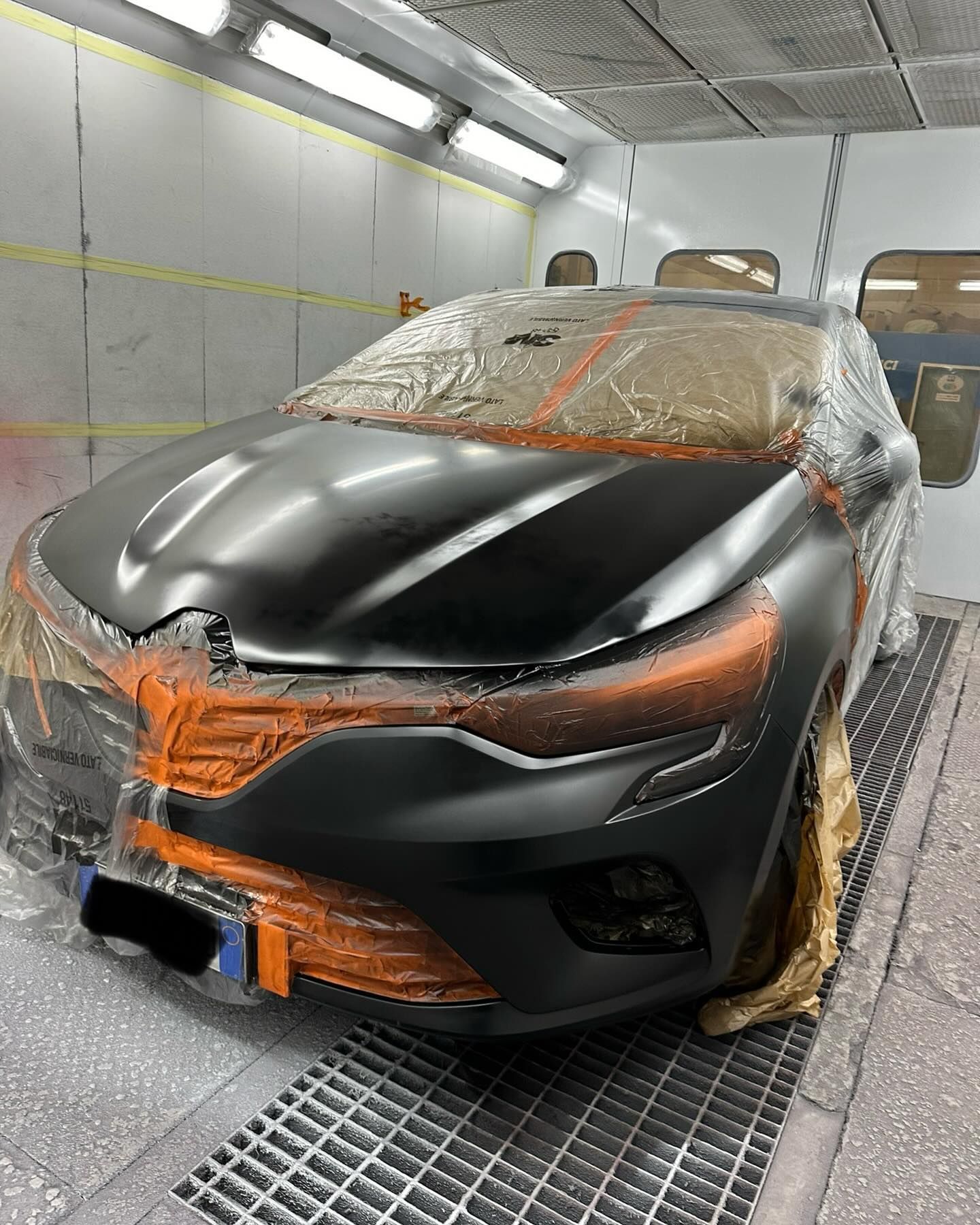 Auto nera mascherata per la verniciatura in una cabina di verniciatura, con nastro protettivo arancione e pellicola di plastica.