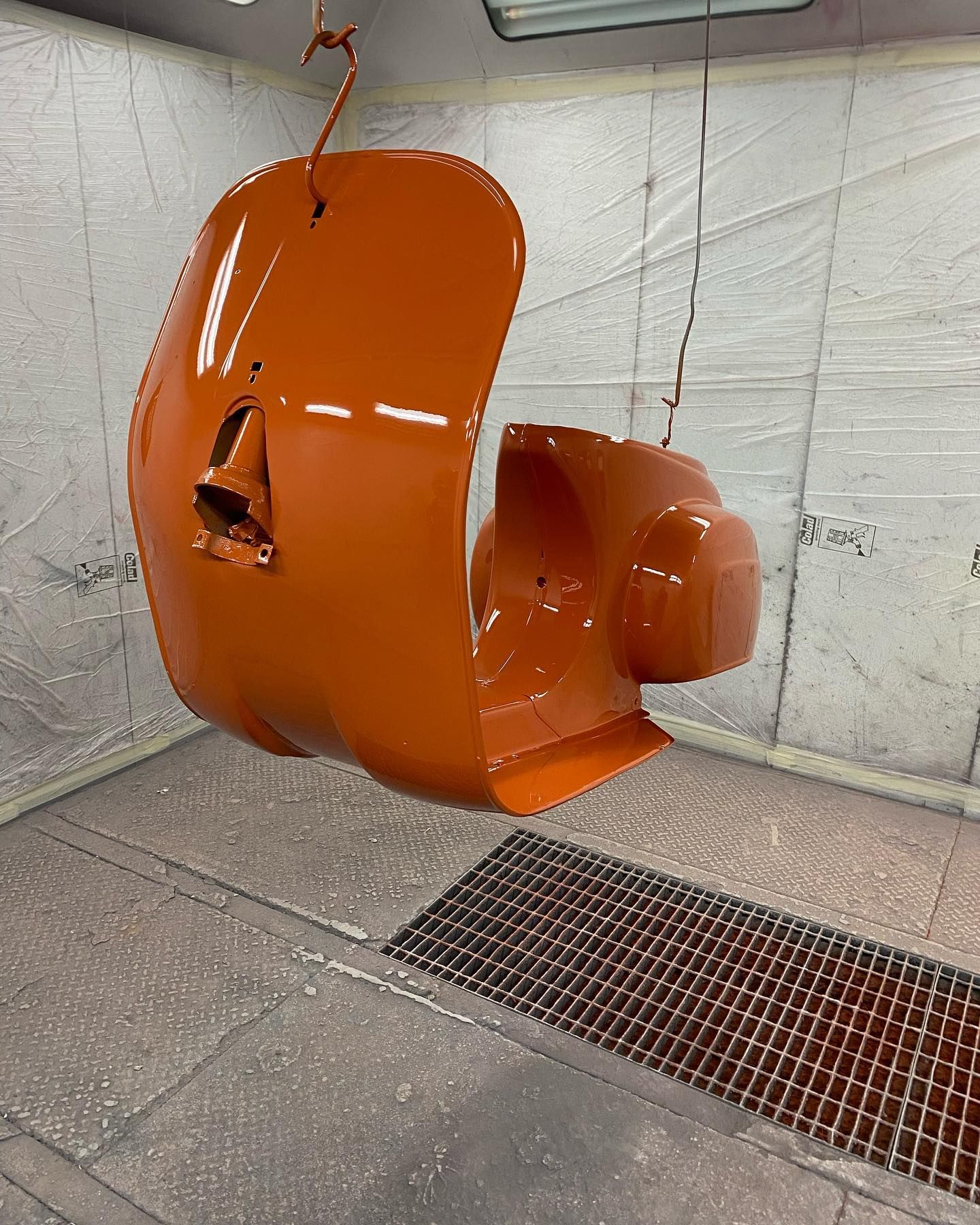 Pezzo di veicolo arancione appeso in una cabina di verniciatura, appena verniciato e lucido.