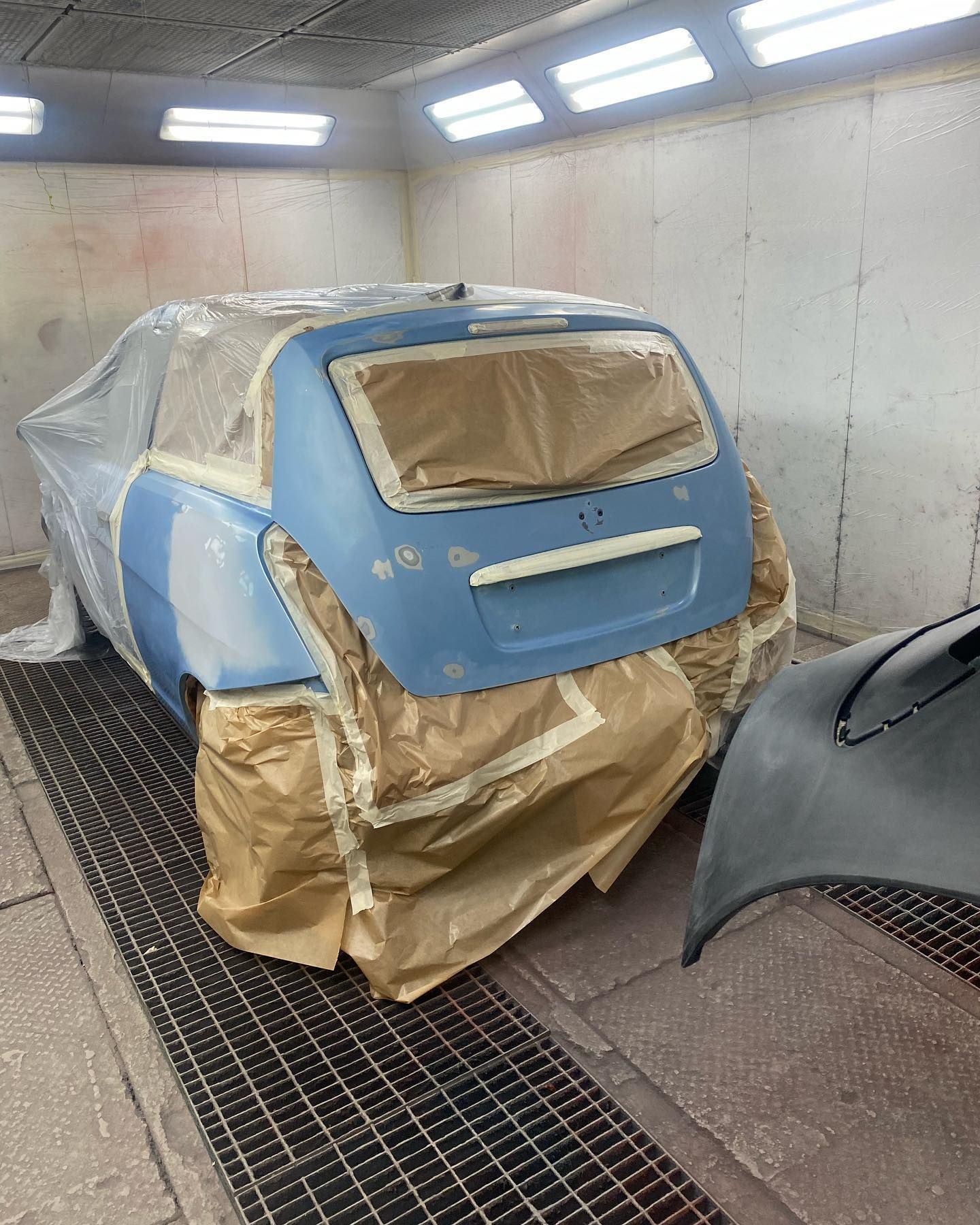 Auto in fase di verniciatura in cabina. La parte posteriore è blu, protetta con nastro adesivo. Il cofano grigio è lì vicino.