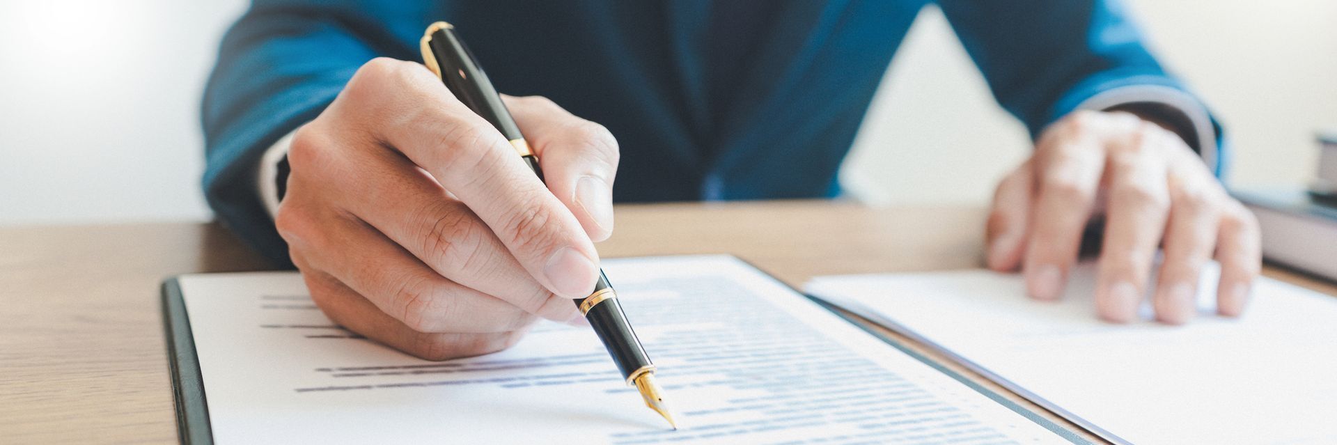 La mano di una persona che tiene una penna mentre scrive un documento su una scrivania di legno.