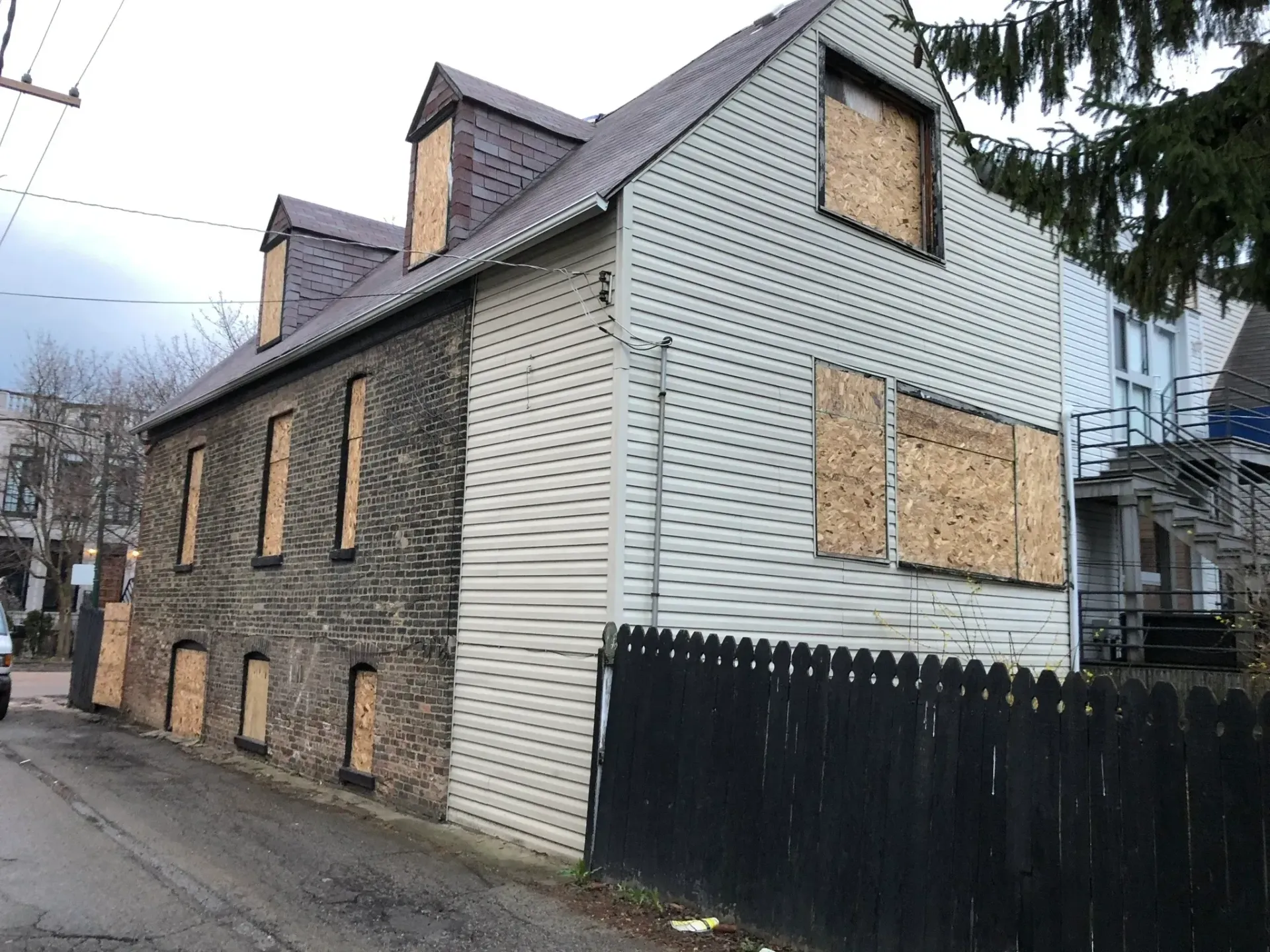 Boarded-up two-story house with dark brick on the left and white siding on the right, dark fence in front.