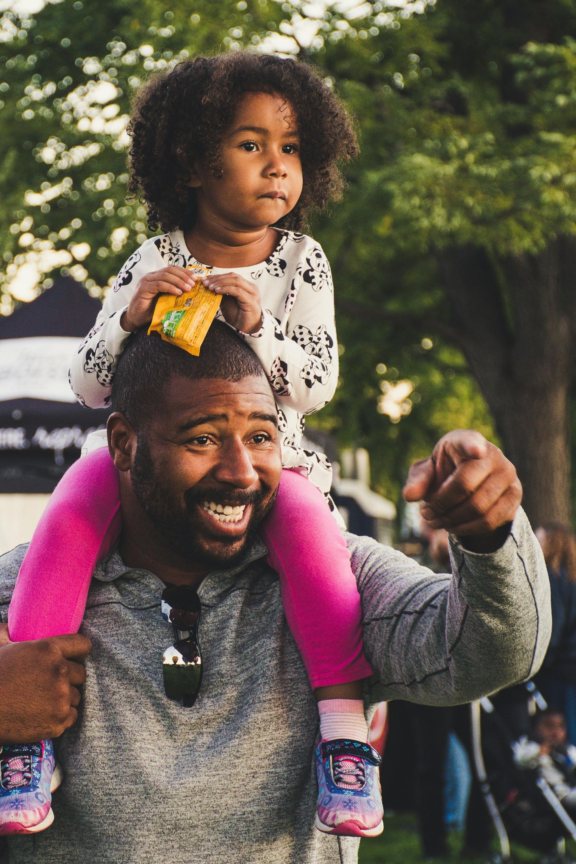 Man smiles, carrying a child on his shoulders outdoors. Child has a snack on their head, points ahead.