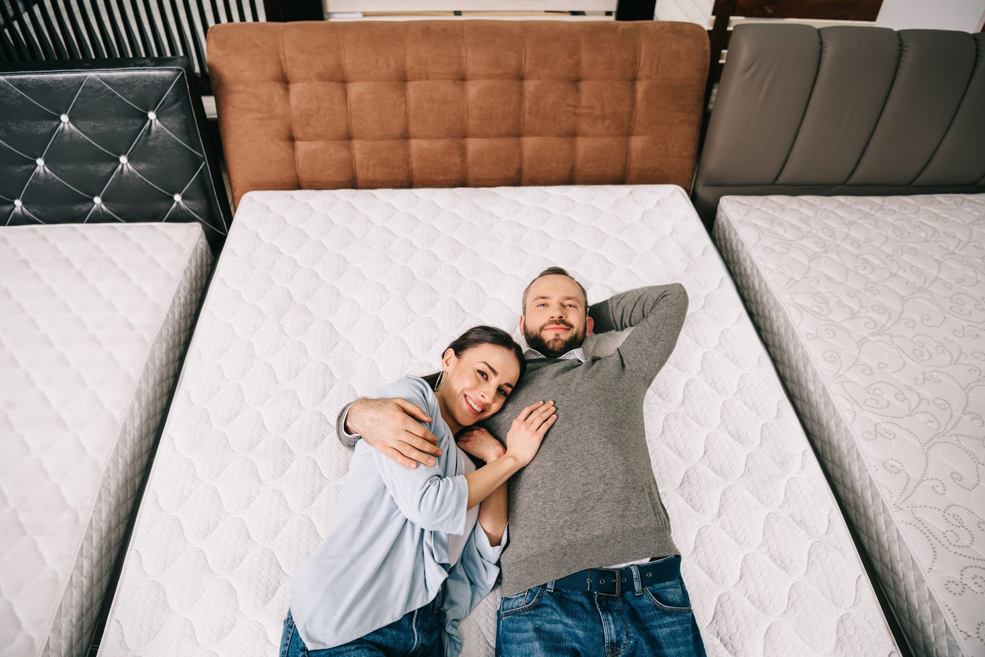 Overhead view of a smiling couple lying on a bed in a furniture store with mattress sets.