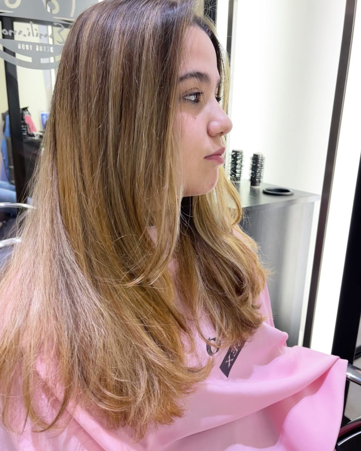 Una persona con cabello castaño, capeado y con mechas, hasta los hombros, en un salón de belleza, vistiendo una capa rosa.