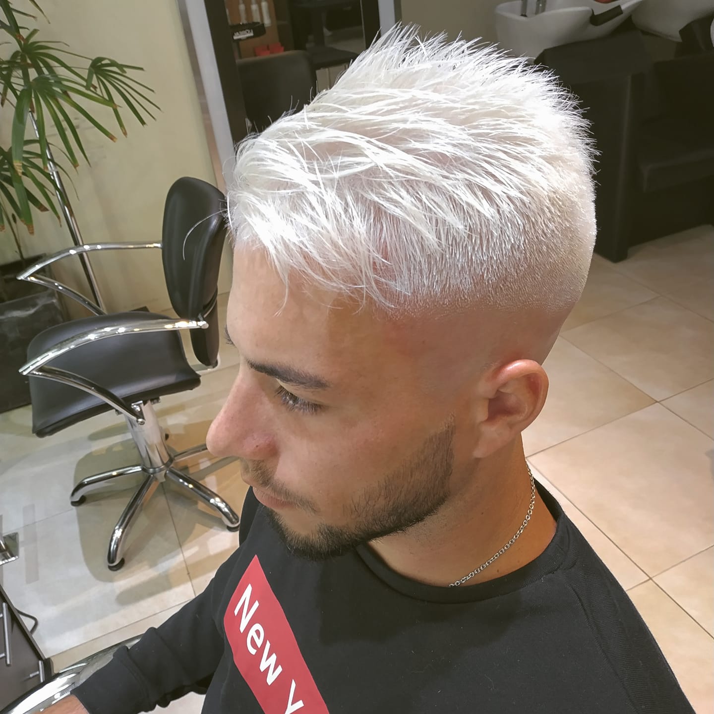 Una persona con cabello corto, blanco hielo y texturizado, con un corte degradado, vista de perfil en un salón de belleza.