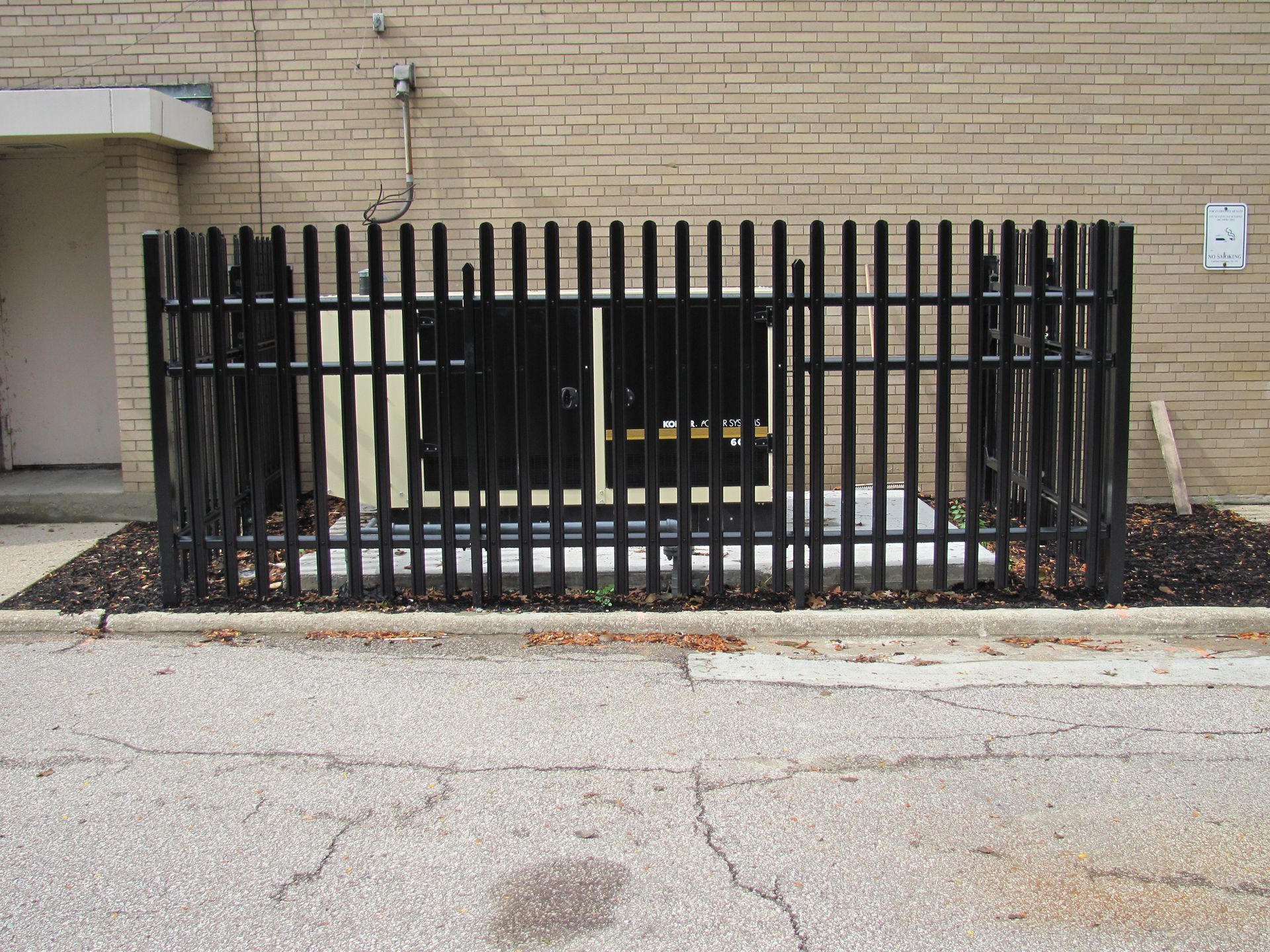 Black metal fence encloses a large, black utility box against a brick building.
