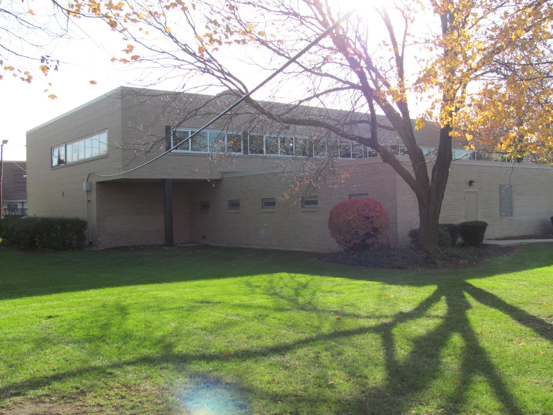 A one-story brick building on a green lawn under a tree with fall leaves. Shadows cover the grass.