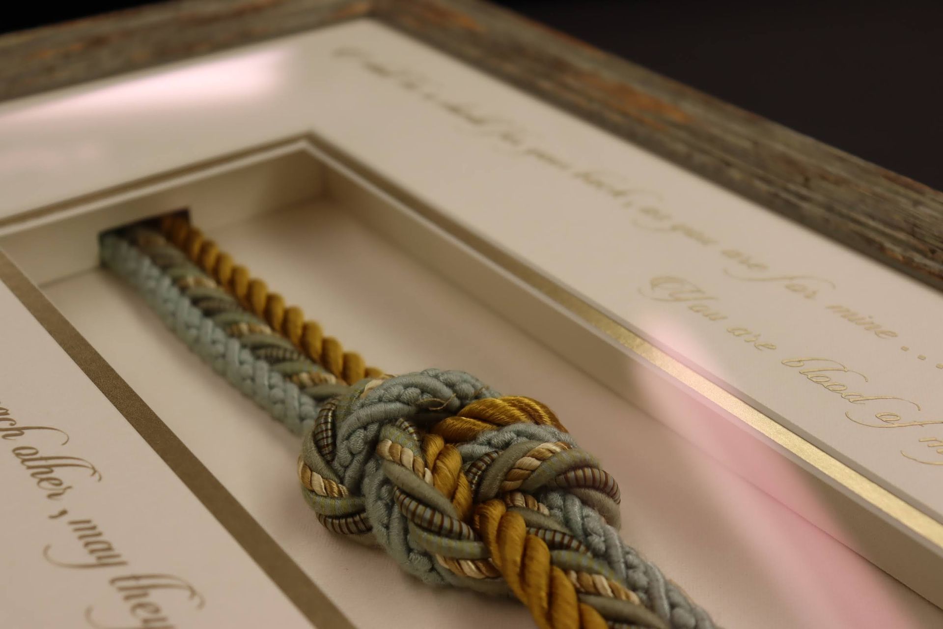 A framed shadow box displaying two braided ropes—one gold and one sage green—tied together in a decorative knot.