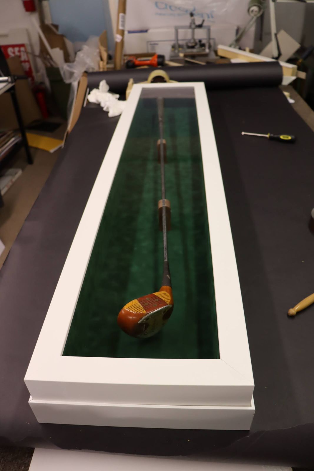 A vintage wooden golf club displayed in a white shadow box with green velvet lining, resting on a black work surface.
