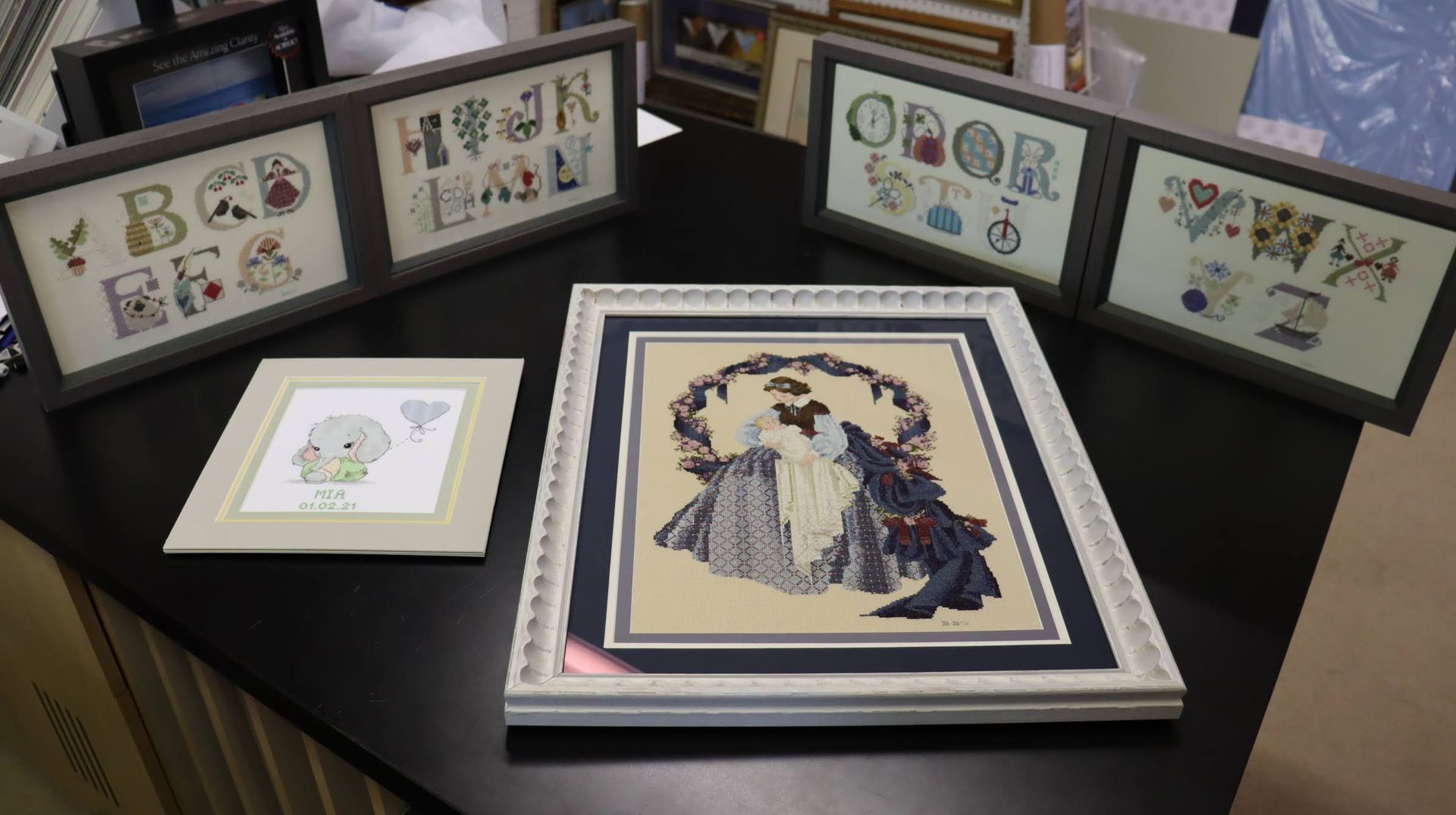 Four framed cross-stitch artworks on a dark surface: an alphabet series in long frames, a small elephant, and a lady.