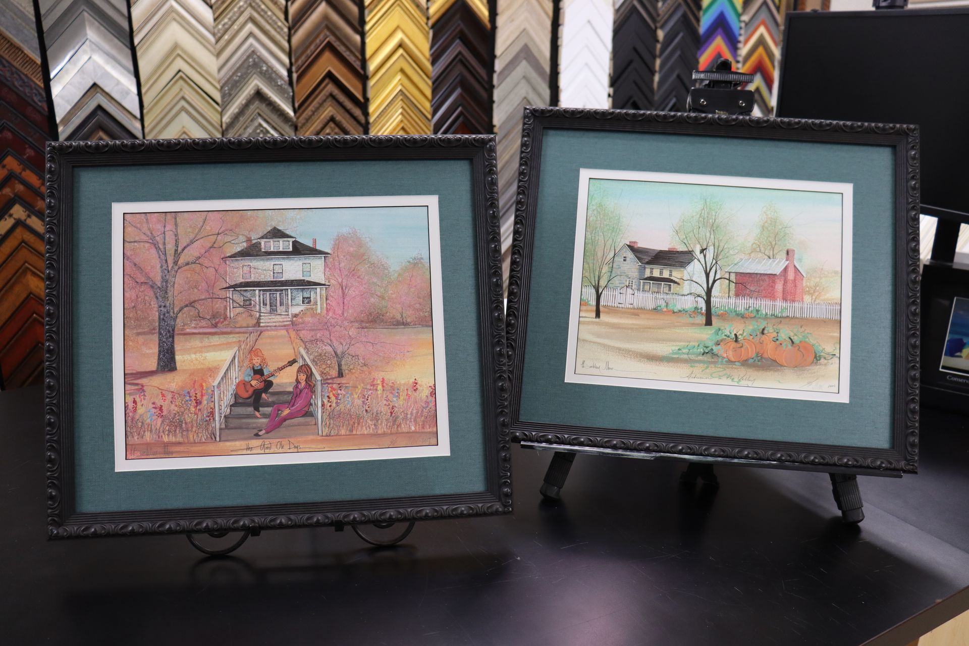Two framed watercolors of houses and landscapes, displayed on easels in front of a wall of various frame samples.