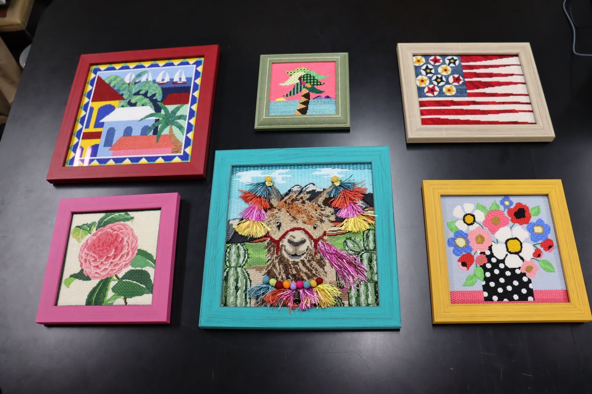 Six colorful framed needlepoint art pieces displayed on a dark surface, featuring varied designs like flowers and a llama.