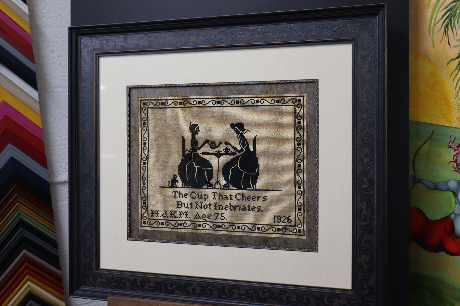 Framed needlework sampler featuring two figures at a tea table with the text 