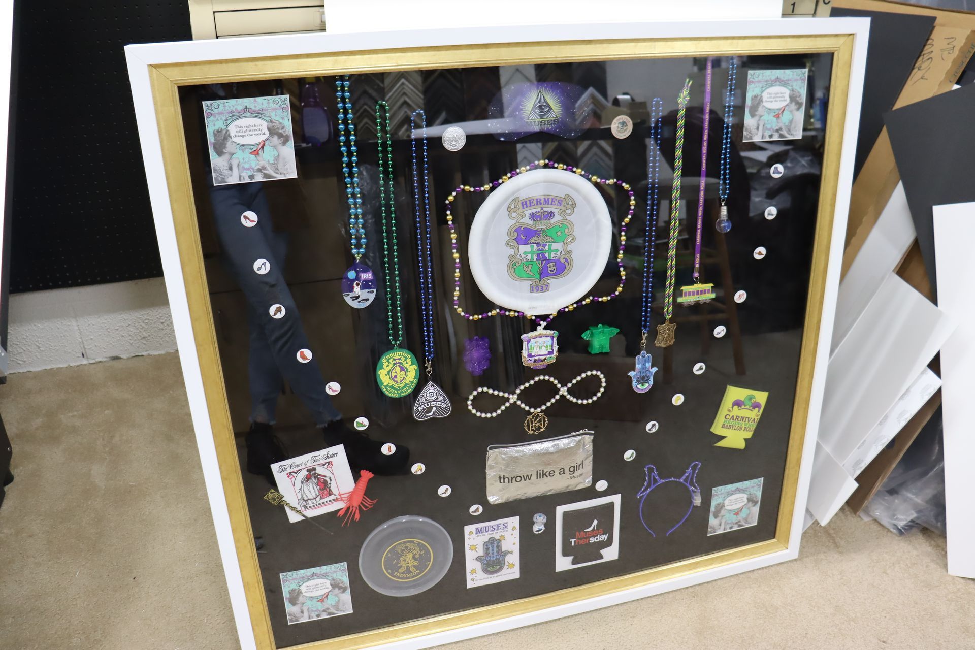 A shadow box display featuring Mardi Gras beads, a circular embroidered emblem, small cards, and tokens on a black backing.