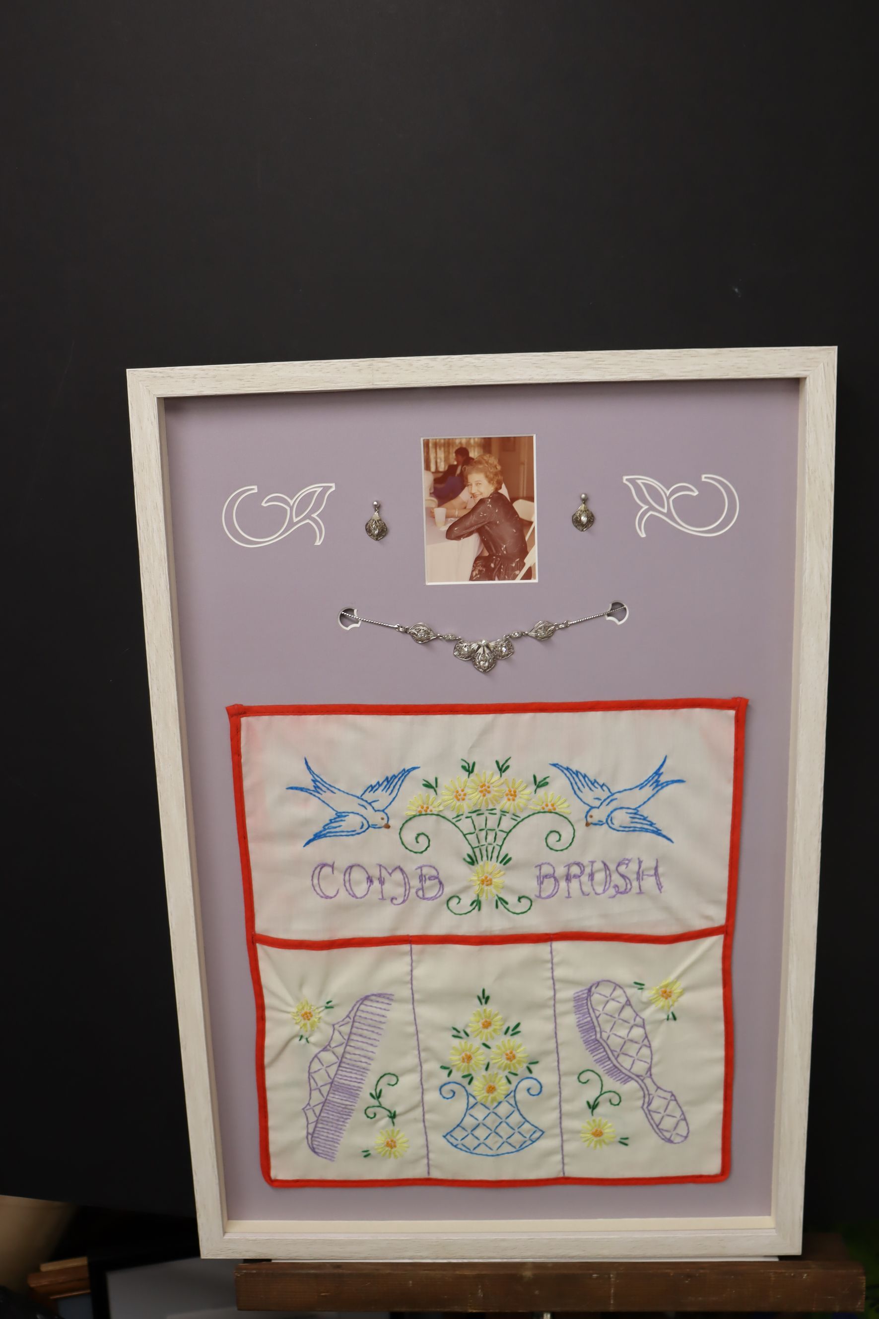 A framed shadow box on a light purple background with a vintage photo, metallic accents, and embroidered fabric panels.