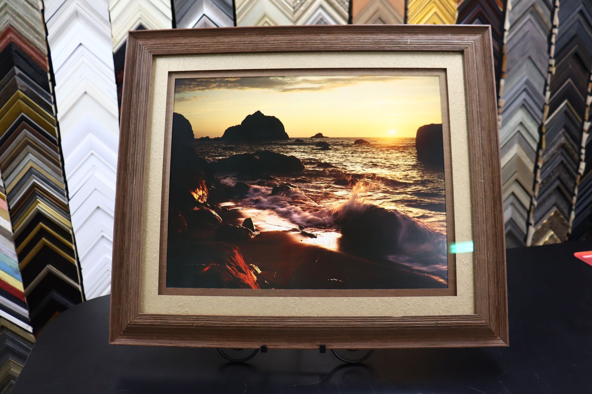 A framed photograph of a rocky coastal sunset with golden light reflecting on the water, displayed in a frame shop.