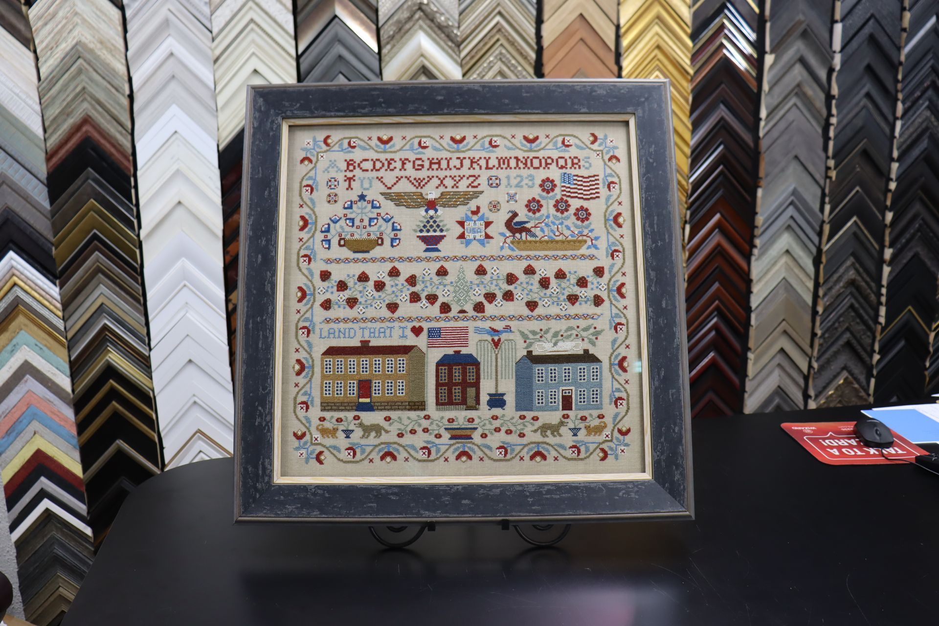 A framed Americana-themed cross-stitch sampler featuring an alphabet, eagle, and buildings, set in a frame shop.