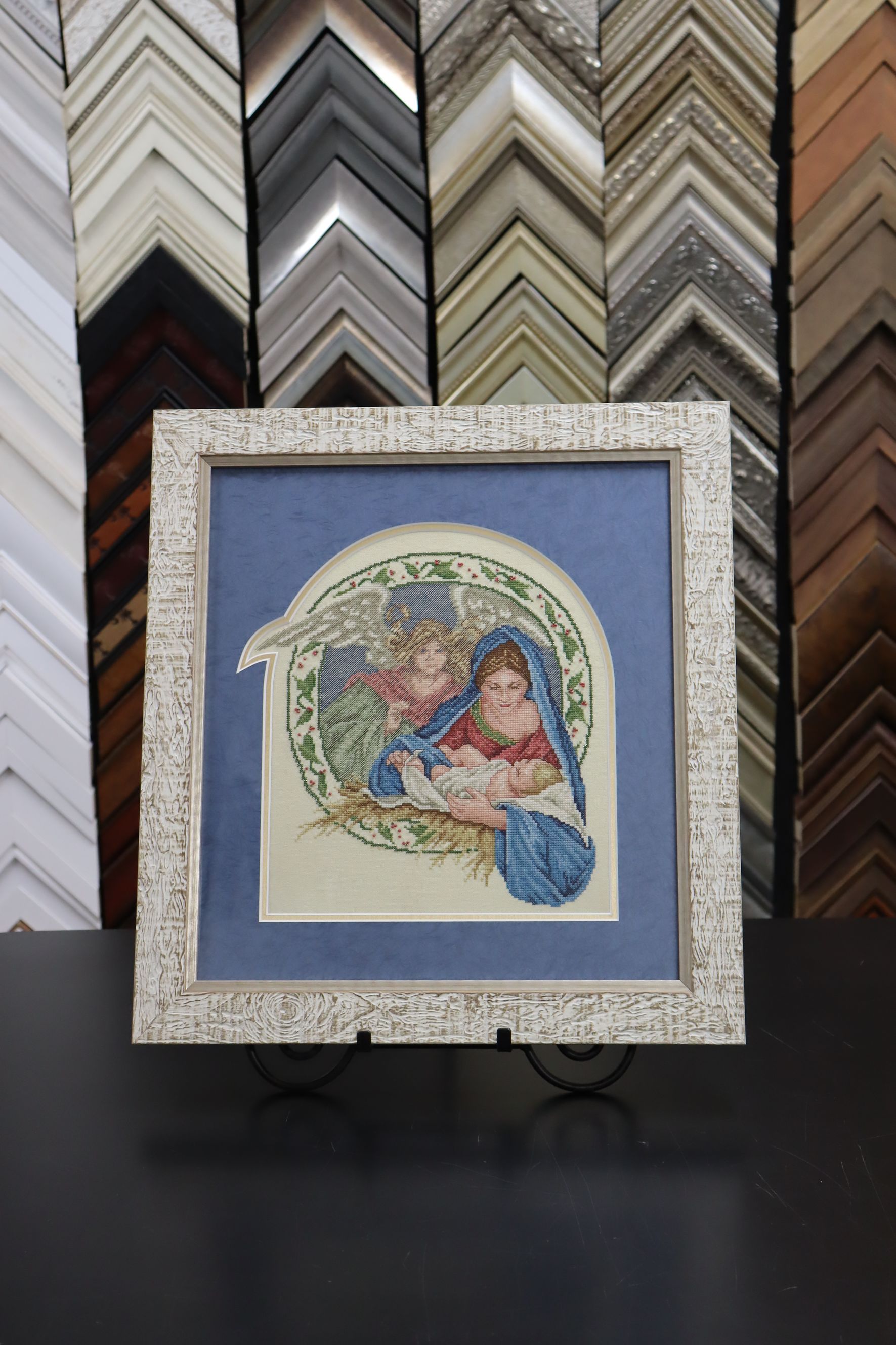 A framed needlepoint of a figure cradling a baby, displayed on a stand in front of various wooden picture frame moldings.