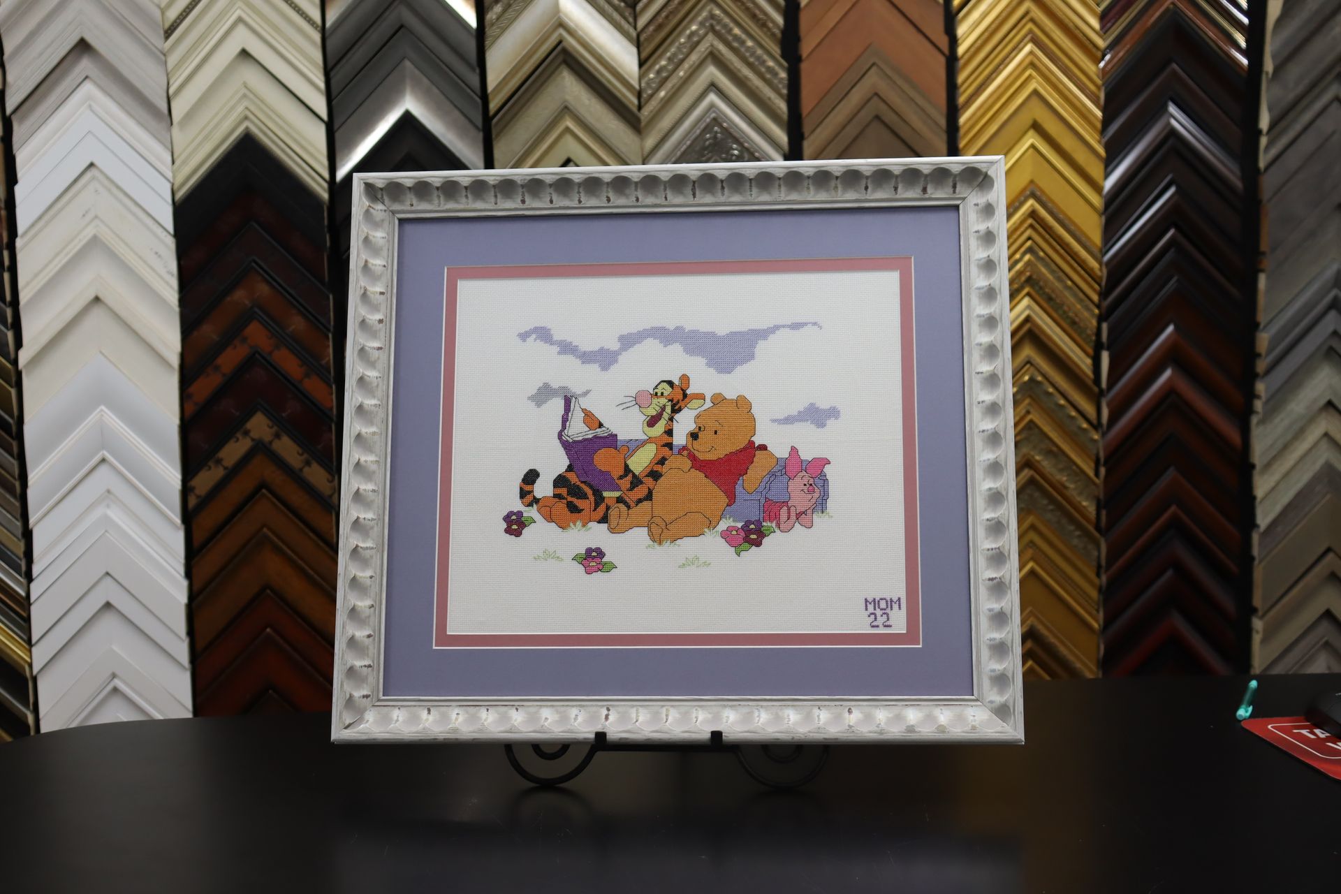 A cross-stitched Winnie the Pooh scene with Tigger, Piglet, and Eeyore in a white ornate frame against wall samples.