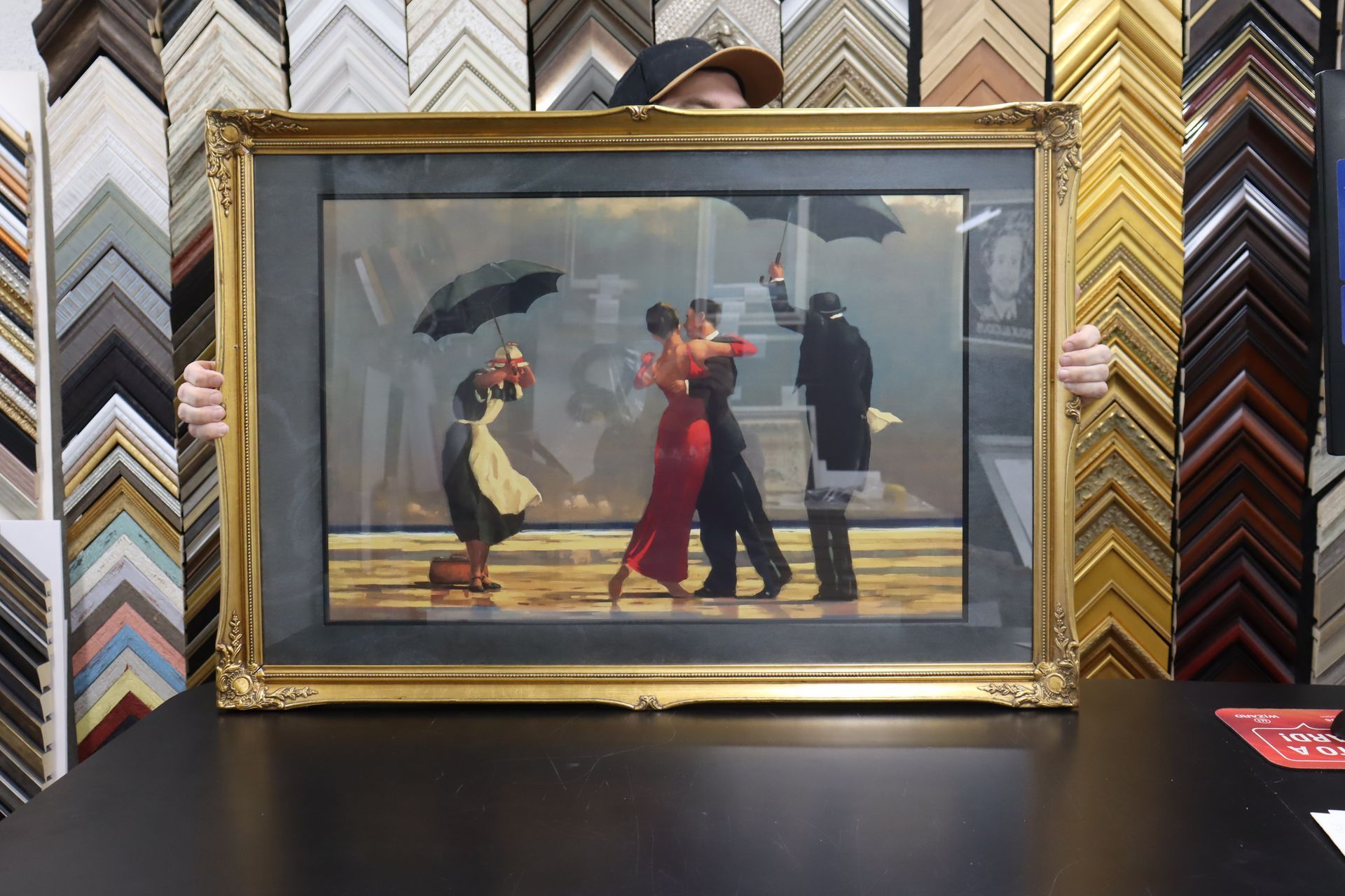 A person holds a framed painting of a couple dancing in the rain beneath umbrellas, surrounded by many picture frame samples.
